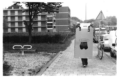 Utrecht, Netherlands, 2000. Black-and-white documentation photograph of a figure in a cardboard box suit walking along a…